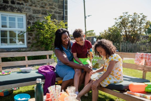 A woman sits with two children outdoors, enjoying a sunny day with snacks and drinks.